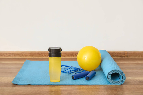 Sports water bottle with skipping rope and ball on mat near light wall - Fotó, kép