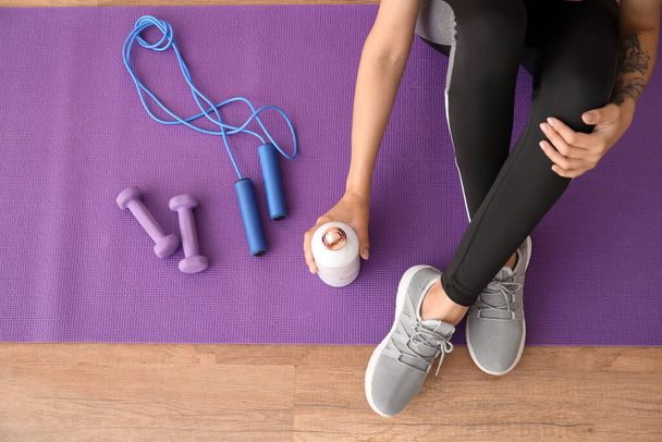 Woman with sports water bottle and equipment sitting on mat in gym - Foto, Imagem