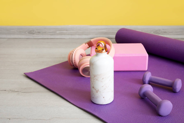 Sports water bottle with equipment and headphones on mat near yellow wall - Foto, Imagem