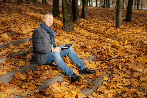 Pensive mature businessman with laptop, sit on steps in golden leaves park. Freelance work and occupation, distance education and electronic sales marketing or trading. Web conference in nature - Foto, imagen
