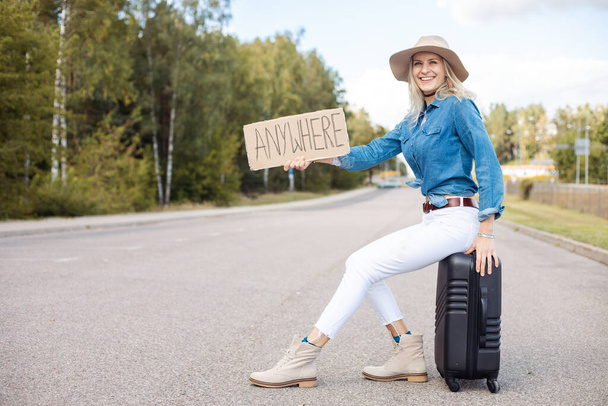 Smiling laughing carefree blond woman in hat and modern jeans shirt hitchhike by roadway with carton nameplate anywhere, sit on baggage suitcase and waiting for car share transportation. Tourist skill - Zdjęcie, obraz