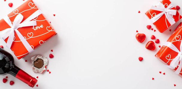 Η ιδέα του Αγίου Βαλεντίνου. Top view φωτογραφία του κόκκινου κουτιά δώρο σε σχήμα καρδιάς καραμέλες σοκολάτα μπουκάλι κρασί και ψεκάζει σε απομονωμένο λευκό φόντο με copyspace - Φωτογραφία, εικόνα