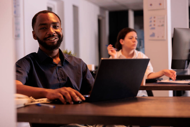 Cheerful employee typing on laptop in coworking space portrait, office manager looking at camera at workplace desk. Smiling african american man using corporate computer app - Photo, image