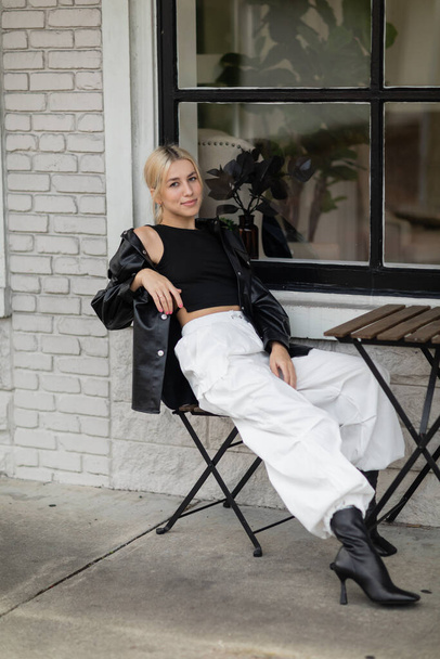 full length of blonde woman in leather shirt jacket and cargo pants sitting in outdoor cafe on street in Miami  - Fotó, kép