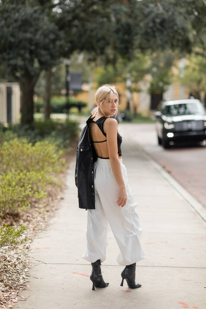 full length of blonde woman in white cargo pants and boots standing with black leather jacket on street in Miami   - Фото, изображение