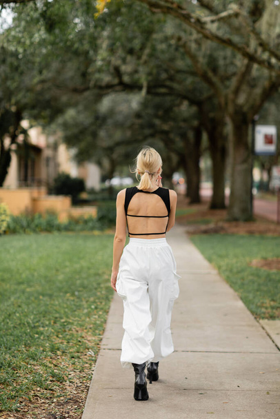 back view of young woman in white cargo pants and black tank top walking on green street in Miami - Foto, Imagen