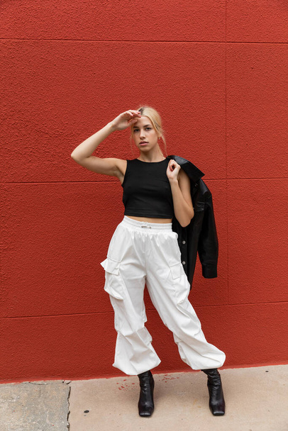 full length of stylish woman in cargo pants standing with leather shirt jacket near red wall in Miami  - Fotoğraf, Görsel