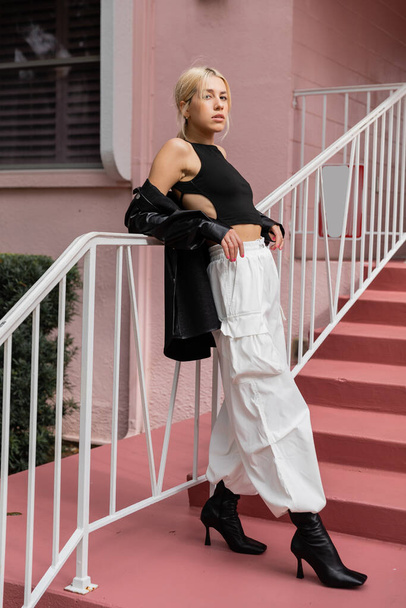 full length of blonde woman in black tank top and cargo pants standing on pink stairs in Miami  - Fotografie, Obrázek