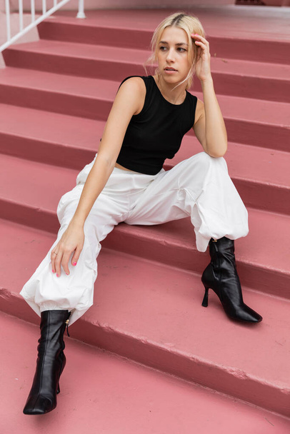 full length of blonde young woman in black tank top and cargo pants sitting on pink stairs in Miami  - Фото, зображення