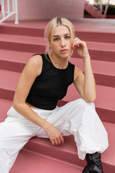 blonde young woman in black tank top and cargo pants sitting on pink stairs in Miami  - Foto, Imagen