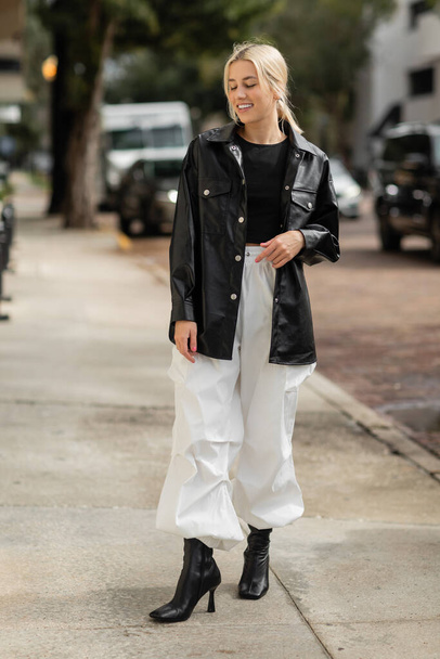 full length of cheerful woman in leather shirt jacket and cargo pants walking on urban street in Miami  - Zdjęcie, obraz