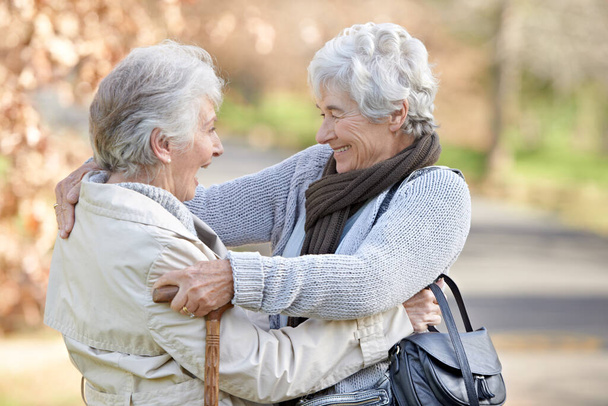 Η φιλία είναι φτιαγμένη για να κρατάει. Δύο ηλικιωμένες κυρίες αγκαλιάζονται με τα φθινοπωρινά σκιασμένα δέντρα στο βάθος - Φωτογραφία, εικόνα