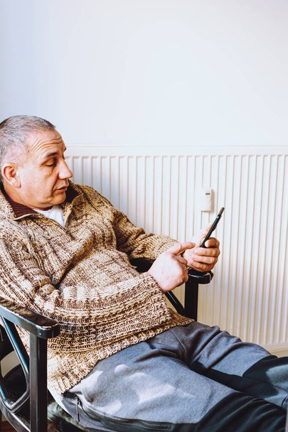 middle-aged man with big fat belly, in warm knitted sweater, sits on chair near heating radiator, rummages through mobile Internet, concept of early aging and depression in middle age men - Foto, Imagen