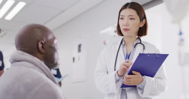 Video of diverse male patient and female doctor with clipboard talking in busy hospital corridor. Hospital, medical and healthcare services. - Filmagem, Vídeo