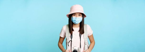 Счастливая азиатская девушка турист, стоя в медицинской маске лица, держа камеру, фотографируя во время отпуска, осмотр достопримечательностей, исследовать за рубежом, стоя на синем фоне. - Фото, изображение