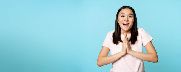 Enthousiaste asiatique femme regarde plein d'espoir, dire s'il vous plaît, montrant geste de mendicité, demandant smth, debout sur fond bleu. - Photo, image