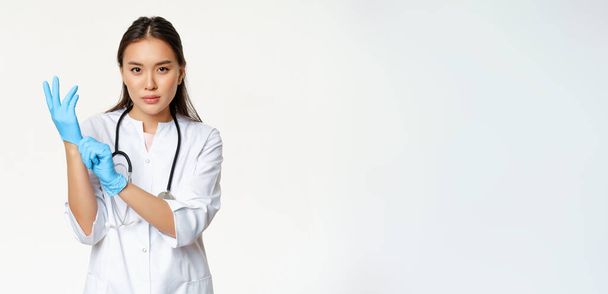 Selbstbewusste Krankenschwester, Ärztin zieht medizinische Gummihandschuhe für die klinische Untersuchung des Patienten an, steht ernst in der Uniform des Gesundheitspersonals, weißer Hintergrund. - Foto, Bild