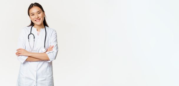 Asian female doctor, physician in medical uniform with stethoscope, cross arms on chest, smiling and looking like professional, white background. - Photo, Image