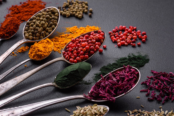 Several types of spices in metal spoons paprika, tomatoes, curry, beets, cumin, turmeric, fennel, spirulina, allspice of different colors on a black concrete background - Foto, Imagem