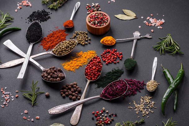 Several types of spices in metal spoons paprika, tomatoes, curry, beets, cumin, turmeric, fennel, spirulina, allspice of different colors on a black concrete background - Foto, Imagen