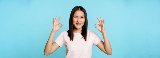 Smiling korean woman winking, showing okay signs, recommending company or store, standing in t-shirt over blue background. - Photo, Image