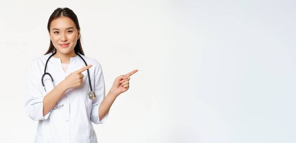 Gesundheitswesen, medizinisches Konzept. Lächelnde asiatische Ärztin, zeigt mit dem Finger nach rechts, zeigt Werbung, Krankenhausinformationen, steht über weißem Hintergrund. - Foto, Bild