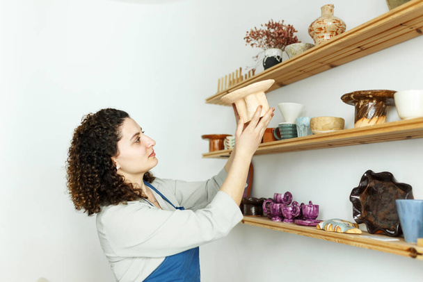 Young girl businesswoman in her workshop is standing next to shelf with ceramic products. Concept small business startup. - Foto, imagen
