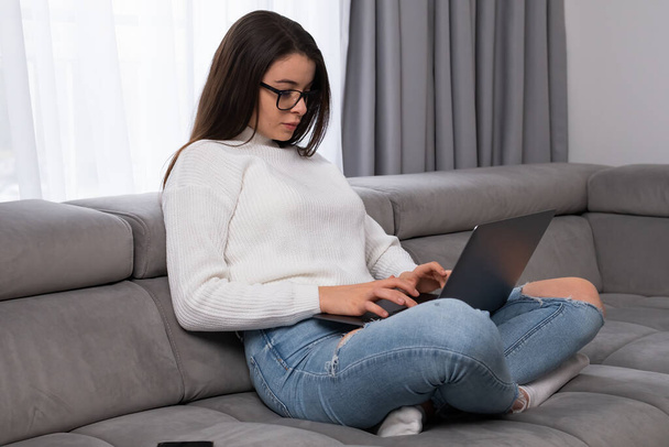 眼鏡の深刻な女性を使用してノートパソコンのチェックメールソファに座っているか、オンラインで勉強. - 写真・画像