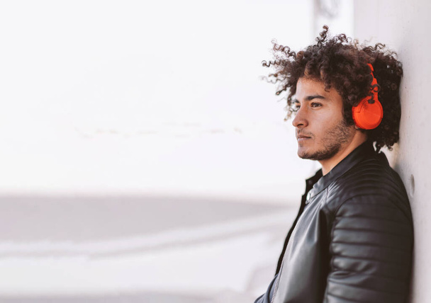 casual pensive young man with curly hair listening to audio track with headphones looking away in daydreaming - Zdjęcie, obraz