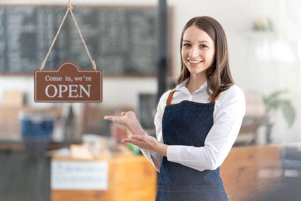 Portrait of a happy waitress standing at restaurant entrance. Portrait of mature business womanattend new customers in her coffee shop. Happy woman owner showing open sign in her small business shop - Fotó, kép