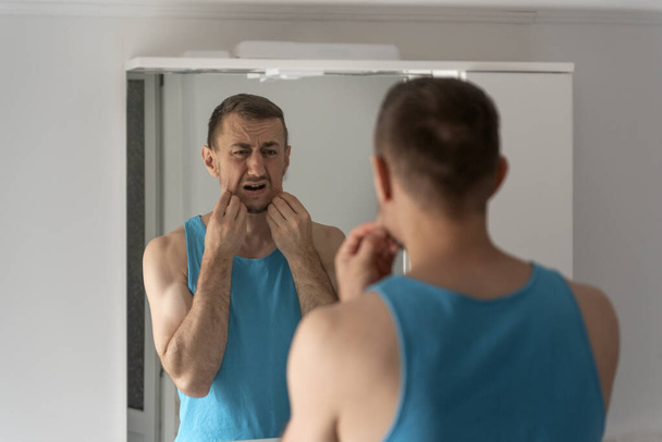Man looks disgruntled at his reflection in mirror in morning in bathroom. Mens facial skin care and shaving - Zdjęcie, obraz