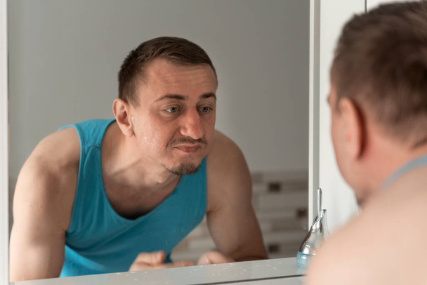 Middle-aged man with no shaving face stands bent over sink and unpleasure looks at reflection in mirror. Morning hygiene. - Fotó, kép