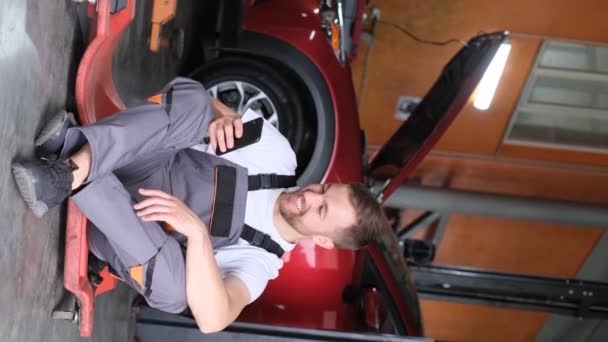 Κάθετη βίντεο, ένας μηχανικός αυτοκινήτων που μιλάει σε ένα smartphone με έναν ιδιοκτήτη αυτοκινήτου. Σχέδιο επισκευής ηλεκτρικών οχημάτων. Βίντεο 4k - Πλάνα, βίντεο