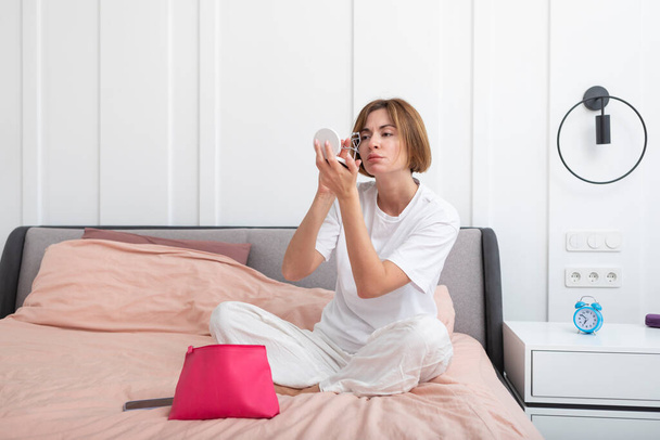 Junge attraktive Frau sitzt auf dem Bett und schminkt sich morgens. Weibliche Morgenroutine und Vorbereitungskonzept - Foto, Bild