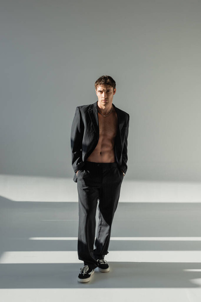 full length of shirtless man in black suit and sneakers standing with hands in pockets on grey background with lighting - Foto, Imagen