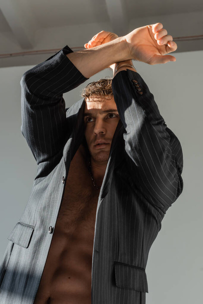 shirtless man in black and striped blazer posing with crossed arms above head on grey background - Fotoğraf, Görsel