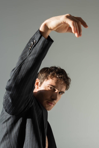 брюнетка мужчина в черном и полосатый блейзер позирует с поднятой рукой, глядя на камеру изолированы на сером - Фото, изображение