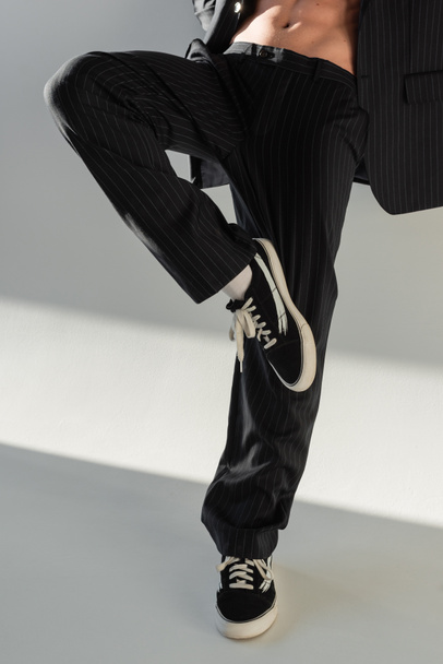 обрезанный вид на модного человека в черных брюках и кроссовках, позирующих на одной ноге на сером фоне - Фото, изображение