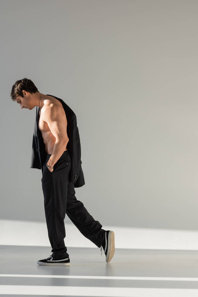 side view of shirtless muscular man in black trousers and sneakers walking with hand in pocket on grey background - Foto, immagini