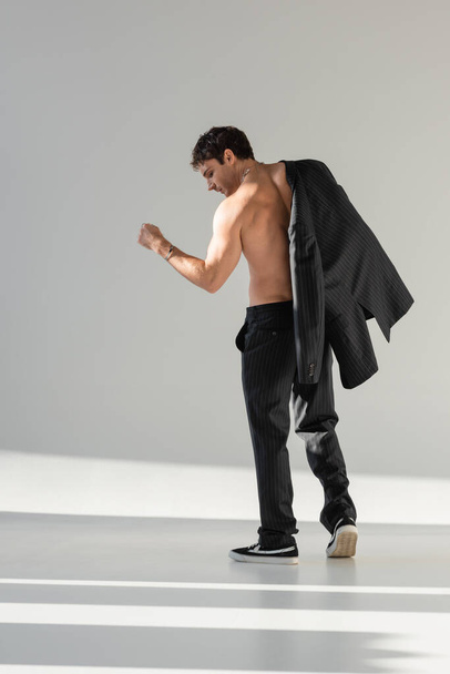 back view of shirtless man in black trousers and sneakers posing with blazer on grey background - Foto, Imagen