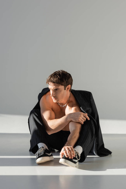 shirtless muscular man in sneakers and pants sitting under black blazer and looking away on grey background - Photo, image