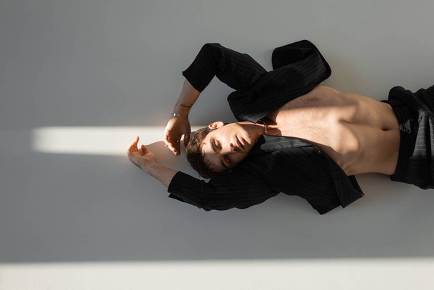top view of shirtless man in black blazer lying with closed eyes in sunlight on grey background - 写真・画像