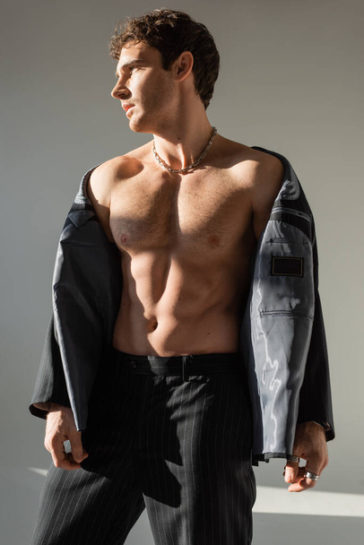 sexy and muscular man looking away while posing in pants and black blazer on shirtless body on grey background - Foto, Bild