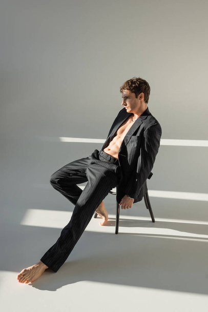 full length of relaxed barefoot man with muscular torso posing in black suit while sitting on chair on grey background - Foto, Imagem