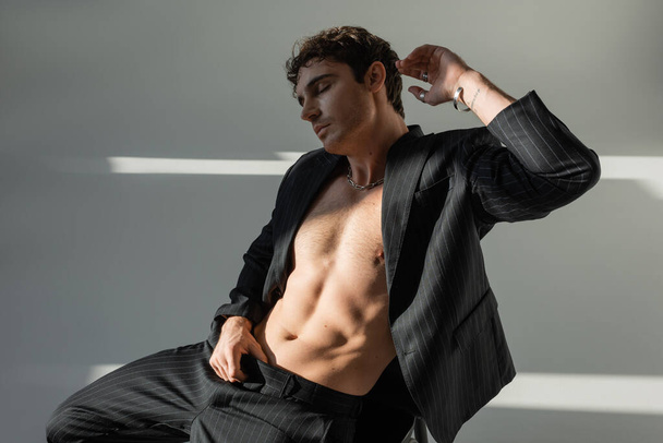 sexy shirtless man in black striped suit sitting with closed eyes and hand near head on grey background - Fotó, kép