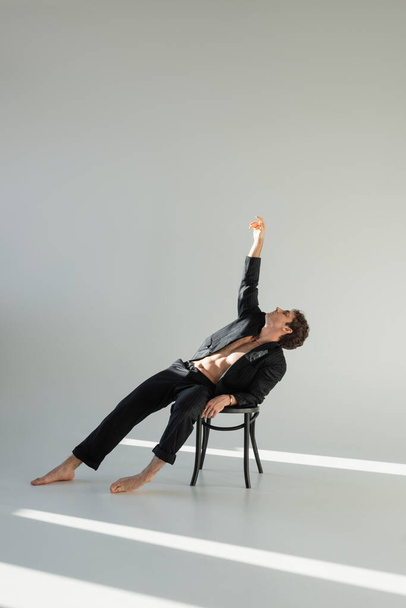 full length of barefoot man in black suit posing on chair with raised hand on grey background with copy space - Fotografie, Obrázek
