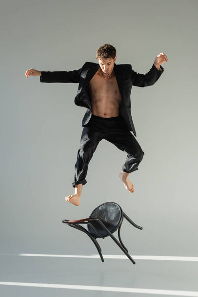полная длина мускулистого человека в черном стильном блейзере, прыгающего со стула на сером фоне - Фото, изображение