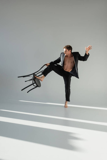 full length of barefoot and shirtless man in black stylish suit posing with chair on grey background - Foto, Imagem