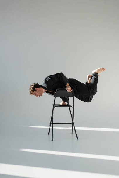 full length of barefoot man in black suit doing trick on chair on grey background - Photo, image