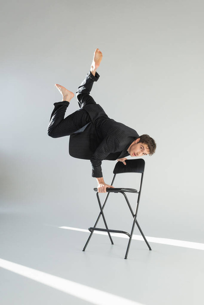 full length of barefoot man in black trendy suit looking at camera and doing handstand on chair on grey background - Foto, Imagem